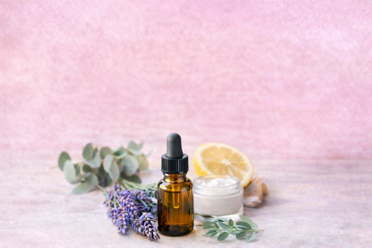 Amberfarbenes Tropffläschchen mit ätherischem Öl vor zartrosa Hintergrund, umgeben von Lavendel, Eukalyptusblättern, halber Zitrone und einer Creme – Symbolbild für ätherische Öle in Naturkosmetik.